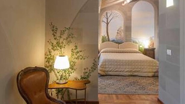 Una habitación con techo de madera, una silla, una lámpara, una mesita y una cama con vista a través de un ventanal arqueado.