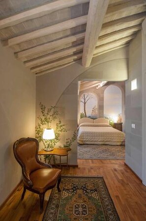 Una habitación con techo de madera, una silla, una lámpara, una mesita y una cama con vista a través de un ventanal arqueado.