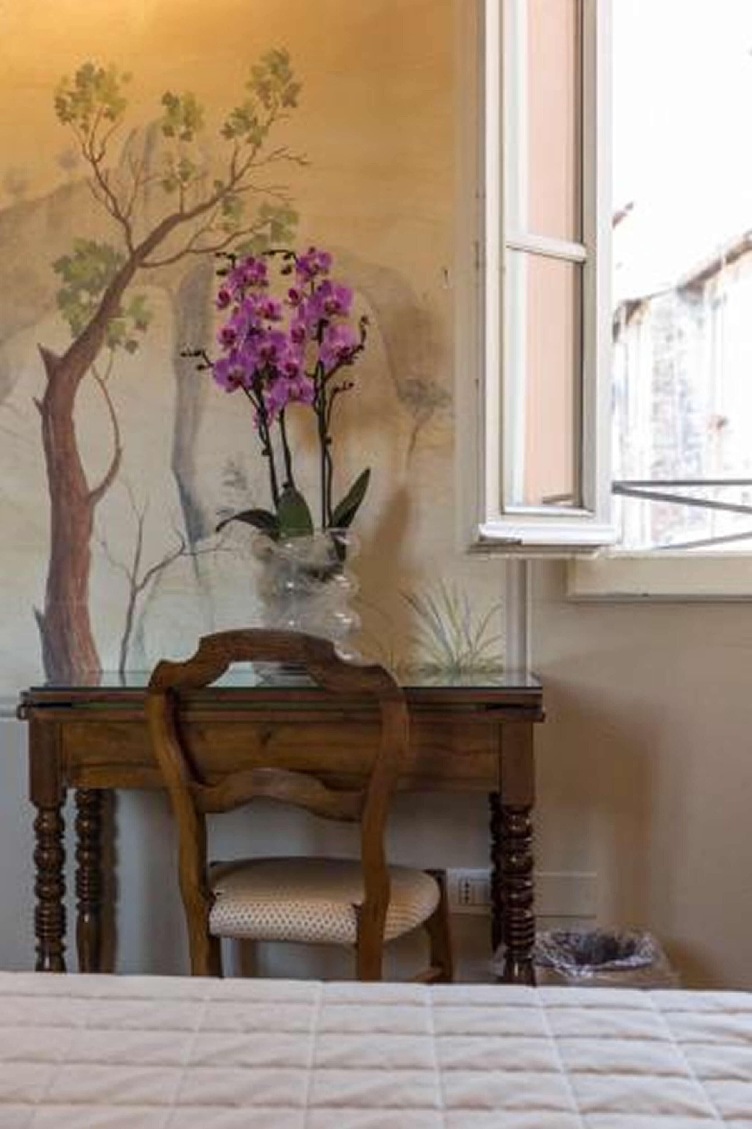 Una camera da letto con un letto, una sedia di legno, un tavolino con un vaso di fiori e un affresco sulla parete.