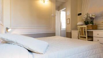 Una cama bien hecha con edredón blanco y almohadas, una mesita de noche con lámpara, una silla de madera y un aparador con espejo y flores.