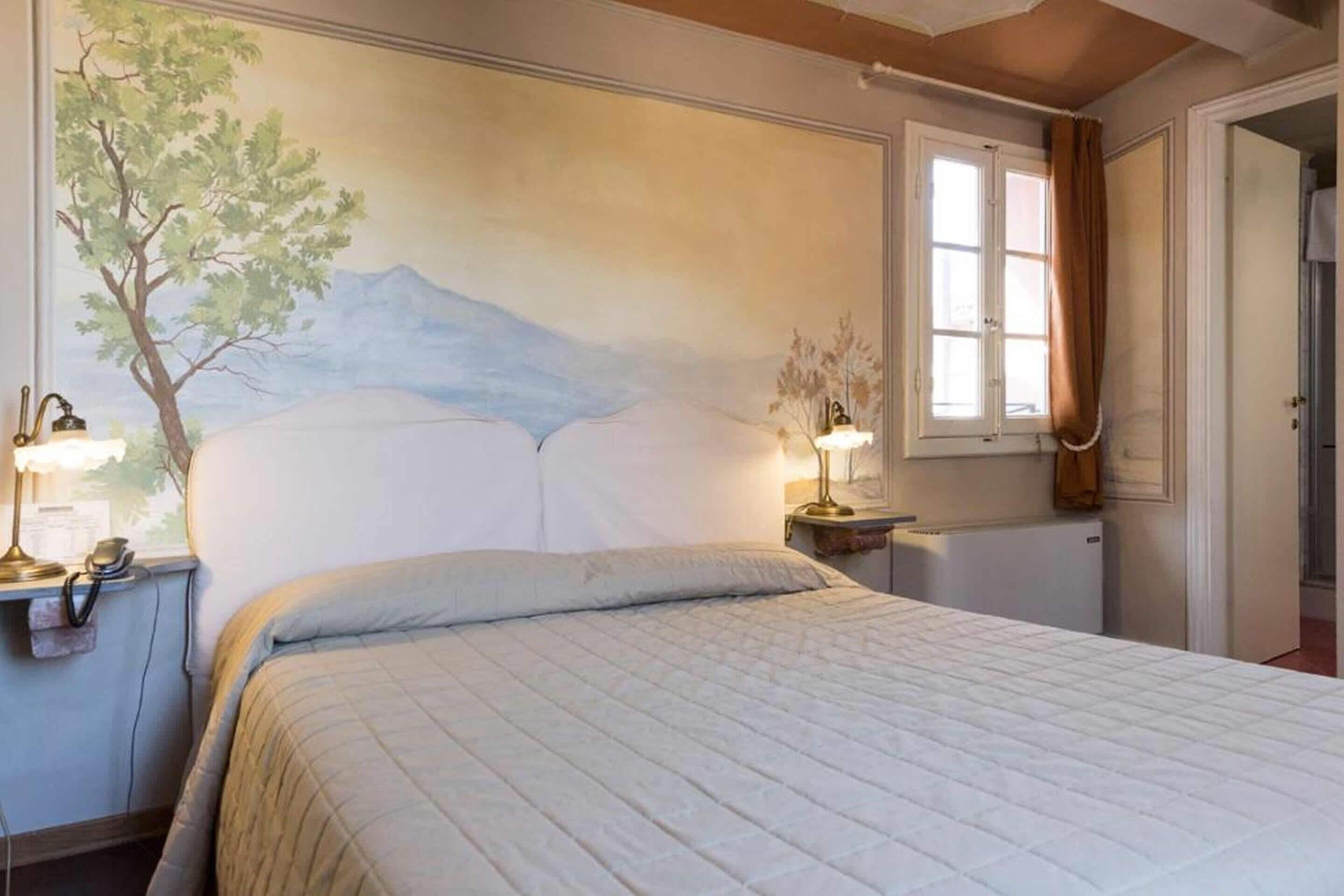 Una camera da letto con un letto, due lampade da comodino, un tavolino, un affresco, una finestra con tende e un termosifone.