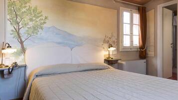 Una camera da letto con un letto, due lampade da comodino, un tavolino, un affresco, una finestra con tende e un termosifone.