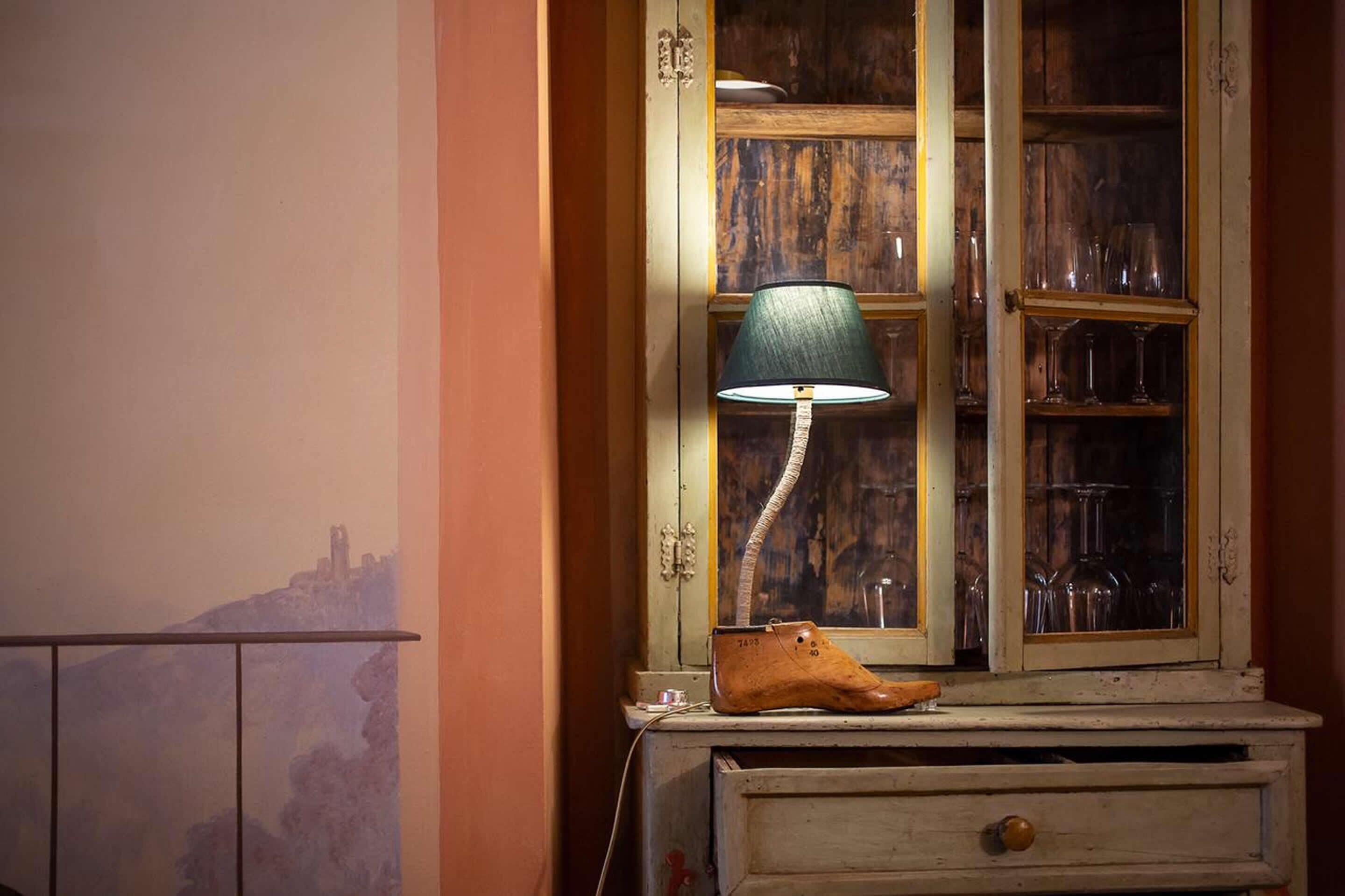 Une lampe ancienne avec un abat-jour vert, une chaussure en bois et un meuble avec des portes en verre contenant des verres.
