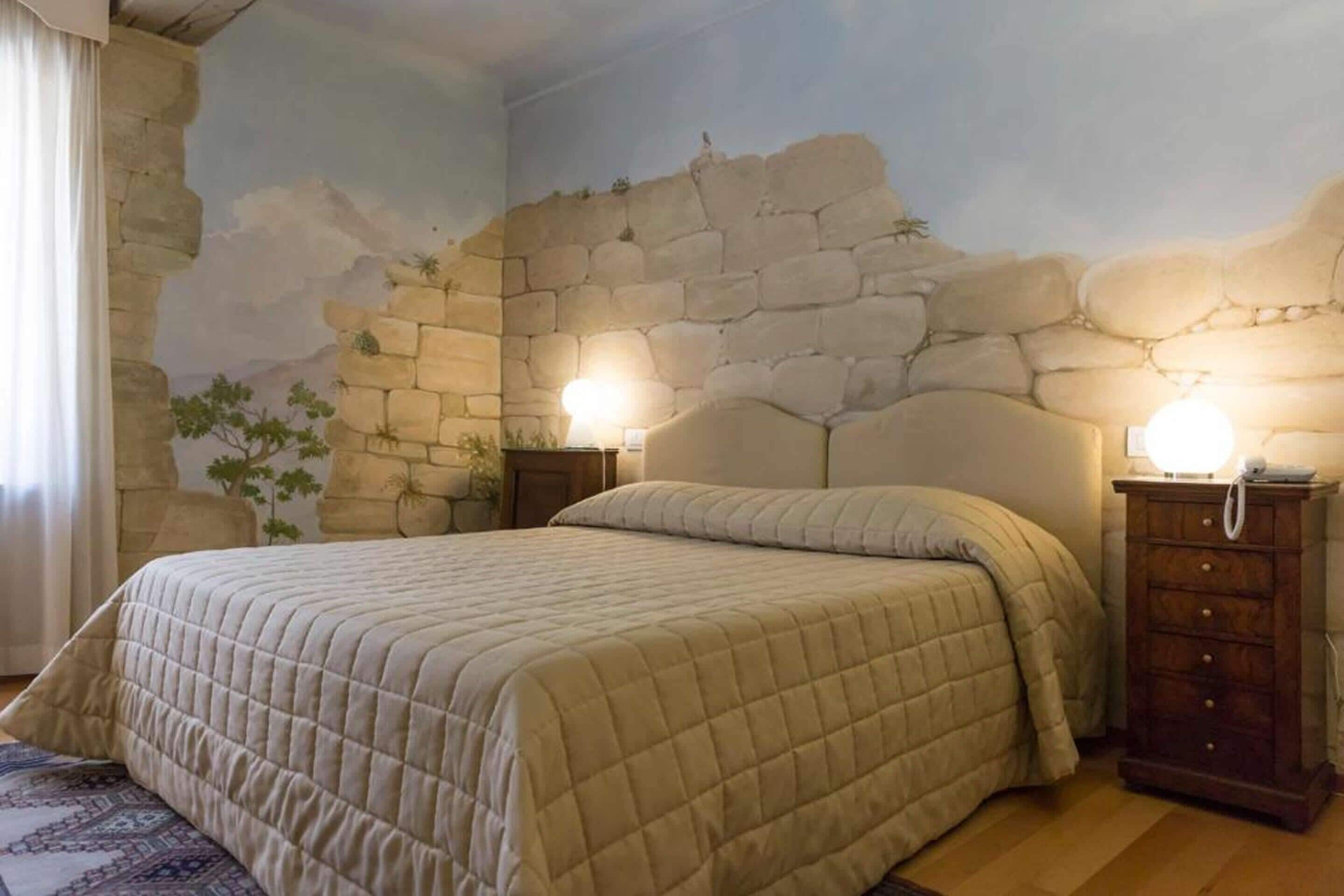 Un dormitorio con un mural de pared de piedra, una cama con colcha color beige, dos mesitas de noche con lámparas y un aparador de madera.