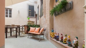 Un estrecho callejón con un banco y una mesa, una fila de figuritas de gnomos en un alféizar, y una jardinera en la pared con follaje.