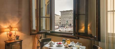 A dining table with tea set and fruit, a window with a view of a historic building, and a lamp on a side table.
