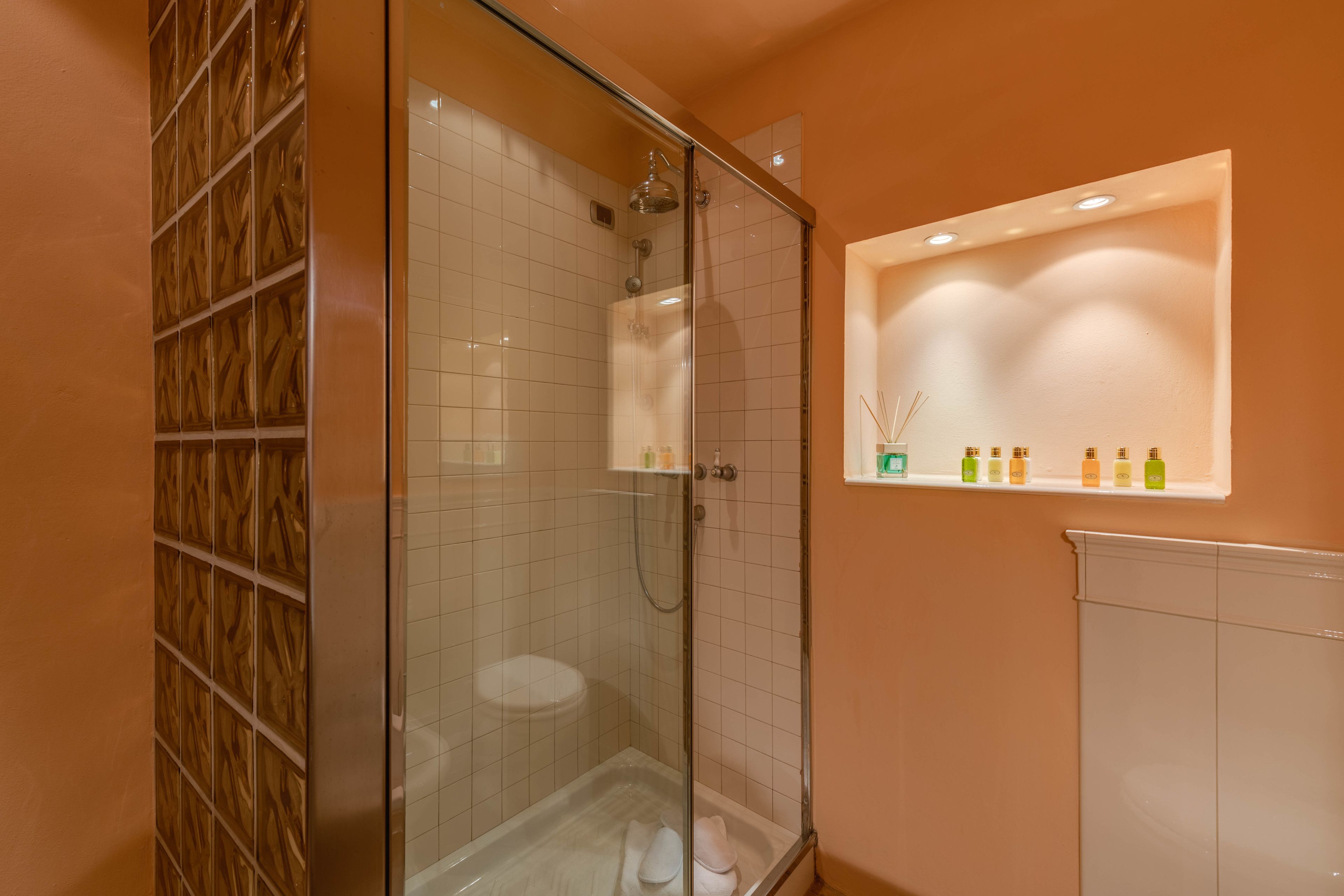 Ducha con cerramiento de vidrio, pared de azulejos blancos, un panel de madera en un lateral y un estante con artículos de aseo.