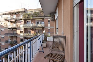 A balcony with a wooden chair, a white railing, and a view of multi-story buildings with balconies. - Abruzzese (Rome)