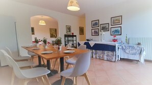 Un comedor con una mesa de madera, sillas blancas y un mantel azul. Hay plantas en macetas sobre la mesa y un aparador blanco al fondo.