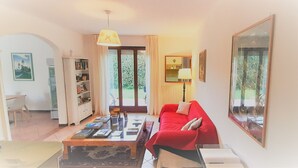 Un salón con un sofá rojo, una mesa de centro con libros, una estantería y un ventanal con cortinas.
