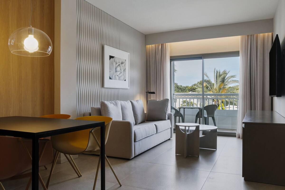 A modern hotel room with a sofa, dining table, chairs, a small balcony, and a large window offering a view of palm trees.