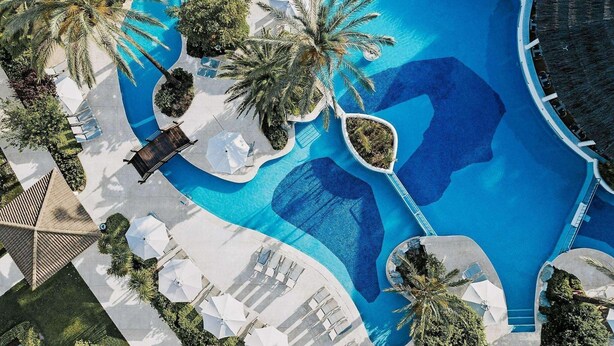 An aerial view of a resort with a large pool, palm trees, and sun loungers. - Prinsotel La Dorada (Muro)