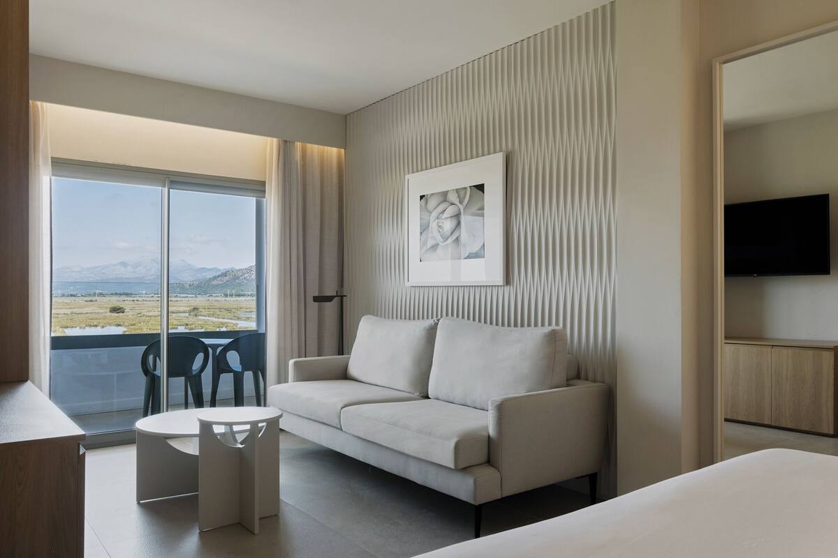 A modern hotel room with a sofa, a small table, a TV, and a large window offering a mountain view.