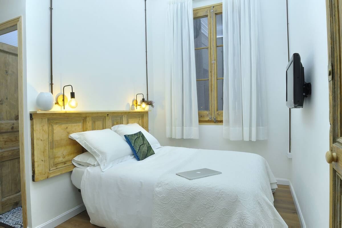 A neatly made bed with a laptop, a wooden headboard, and a wall-mounted TV.