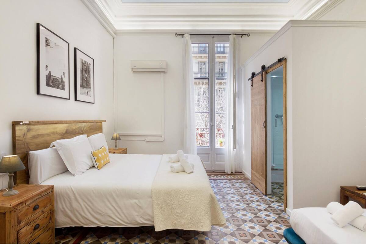 A hotel room with a large bed, a wooden headboard, a nightstand with a lamp, and a view of the outside through a glass door.