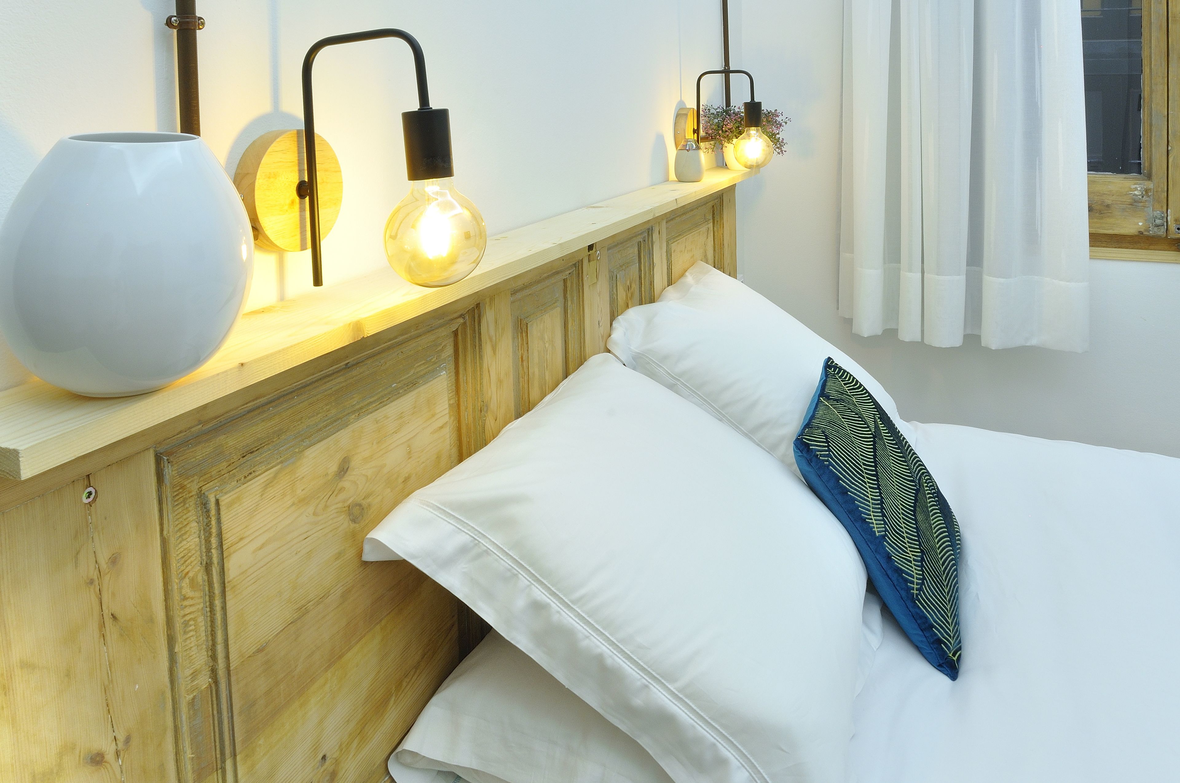 A bedroom with a wooden headboard, white bedding, and a bedside table with a white vase and two wall-mounted light fixtures.