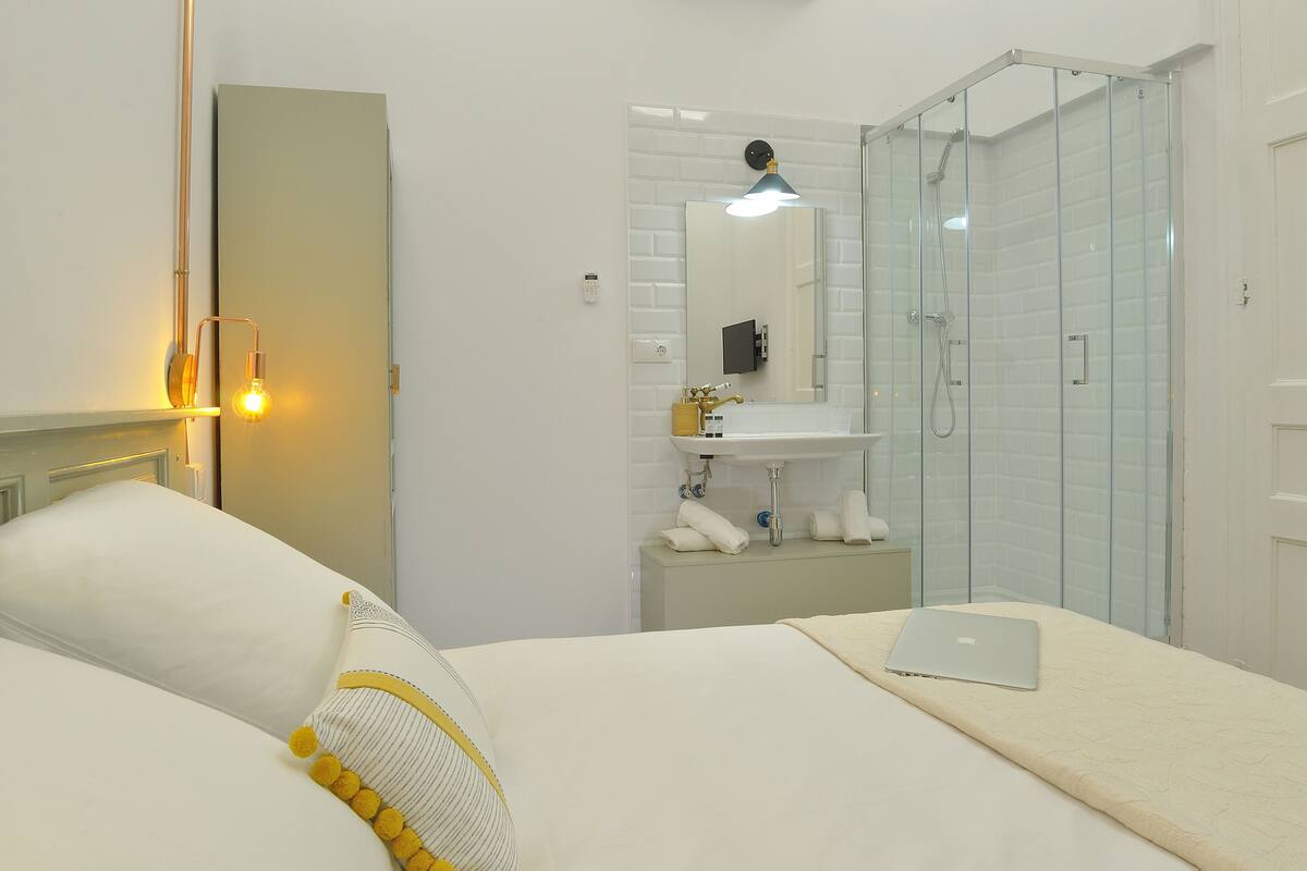 A modern bathroom with a glass shower enclosure, a sink with a mirror, and a bed with a laptop on it.