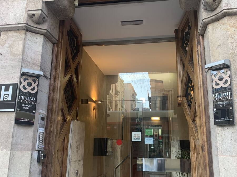 Entrada de hotel con fachada de piedra, puerta de vidrio y marco de madera. Se ven letreros que indican 'Ciudad Condal Boutique Hotel' y un cartel que dice 'Empujar, gracias'.