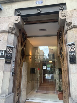 Entrada de hotel con fachada de piedra, puerta de vidrio y marco de madera. Se ven letreros que indican 'Ciudad Condal Boutique Hotel' y un cartel que dice 'Empujar, gracias'.