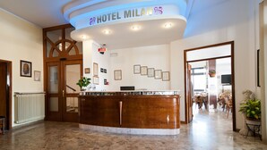 A hotel reception area with a wooden reception desk, framed certificates on the wall, and a sign reading "HOTEL MILAN". - Hotel Milan (Rosolina)