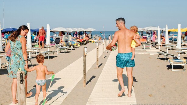 A beach scene with people walking on a boardwalk, beach chairs, umbrellas, and lounge chairs. - Hotel Milan (Rosolina)