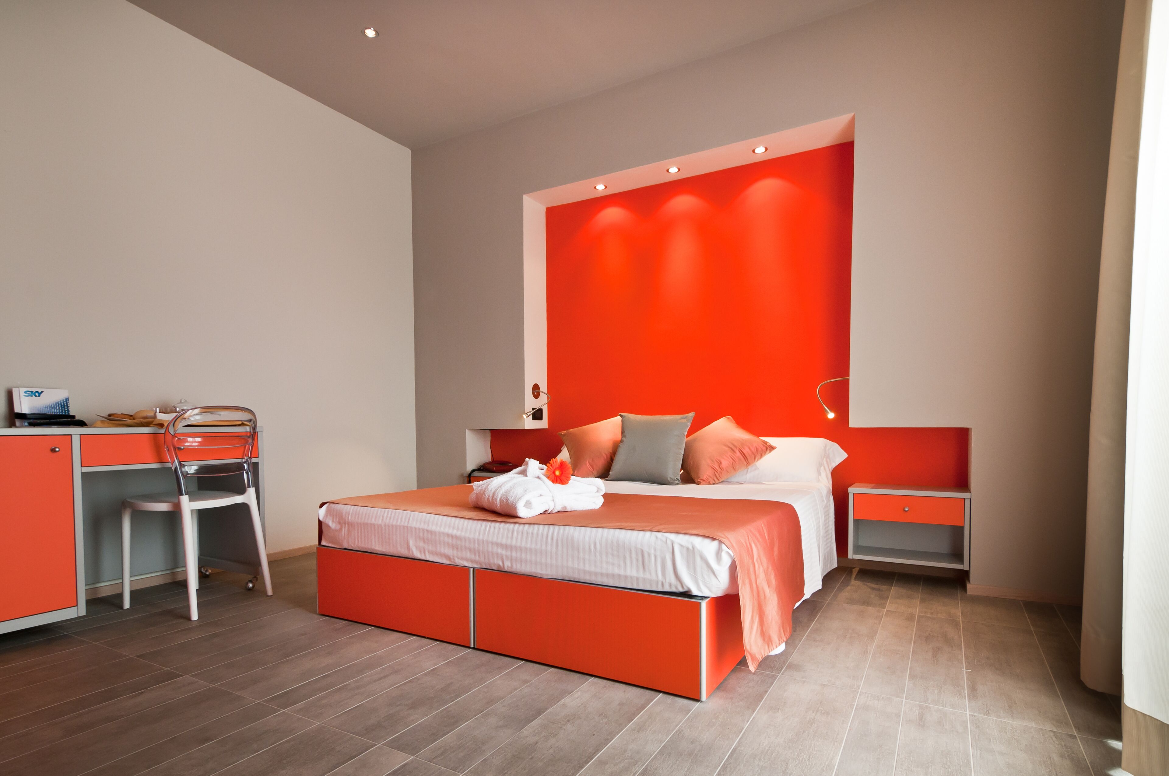 Ein modernes Schlafzimmer mit roter Akzentwand, einem ordentlich bezogenen Bett, einem Schreibtisch mit Stuhl und einem Nachttisch.