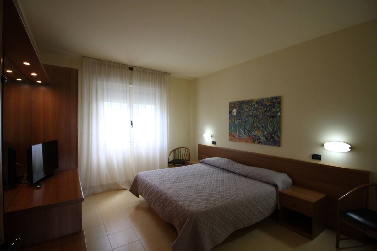 A hotel room with a bed, a desk with a TV, a chair, a nightstand, and a painting on the wall.