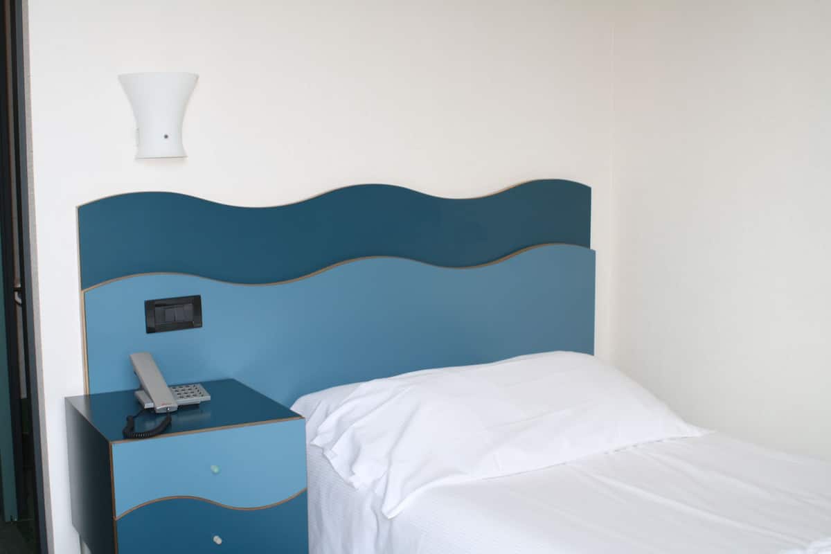Una cama individual con cabecera azul, un teléfono en la mesita de noche y una lámpara fijada a la pared.