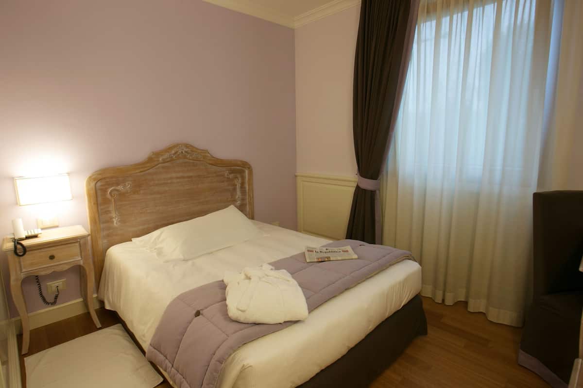 Una cama bien hecha con edredón blanco y manta morada, una lámpara de noche y un teléfono sobre una mesita de noche de madera.