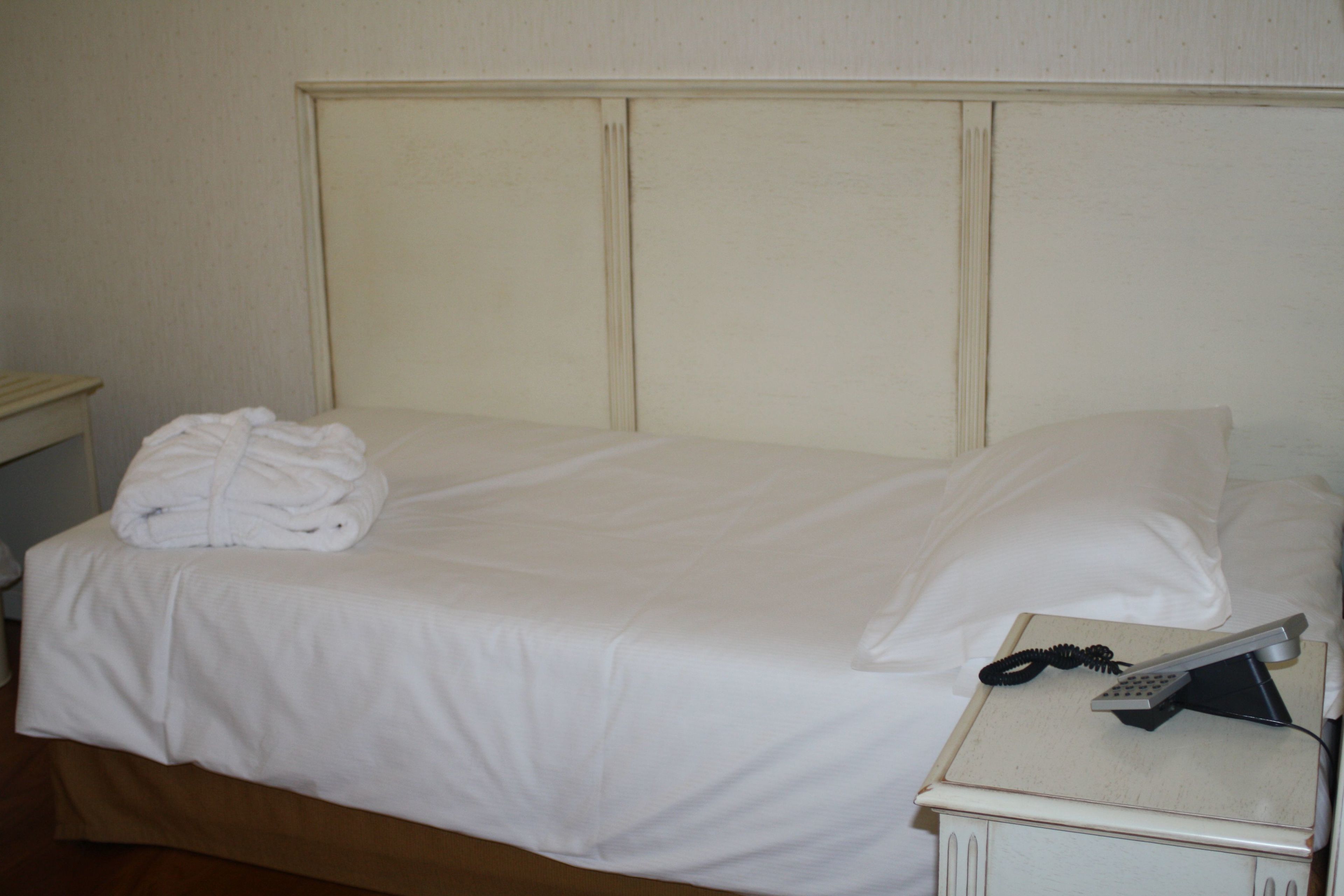 A neatly made bed with white linens, a folded white robe on the side, and a bedside telephone.