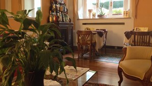 Ein Wohnzimmer mit einem Holzkabinett voller Flaschen, einem Esstisch mit Stühlen und einem großen Fenster mit Vorhängen.