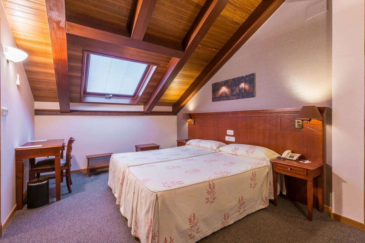 Un dormitorio con techo de madera, tragaluz, cama con ropa de cama floral, escritorio y mesita de noche con teléfono.