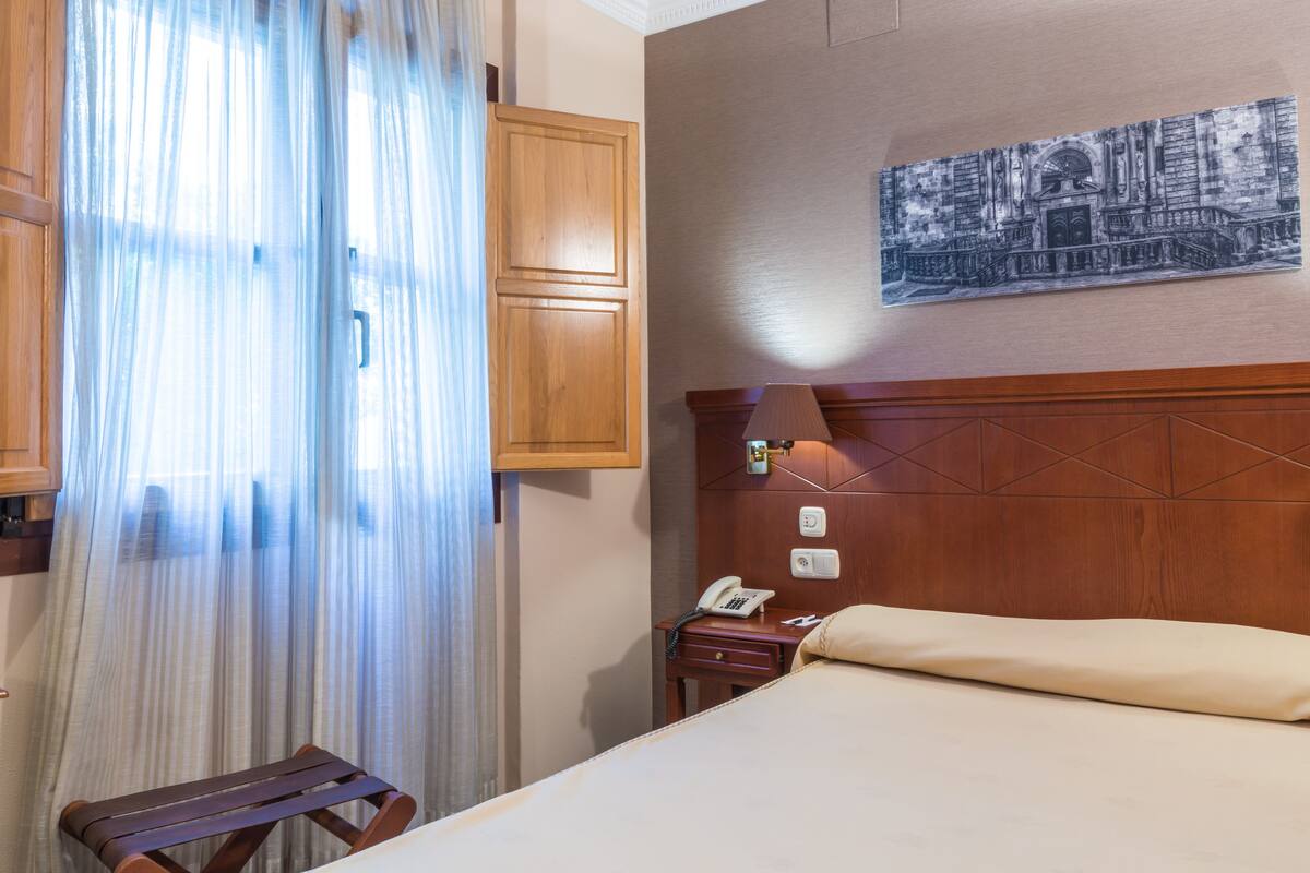 Una habitación de hotel con una cama, una mesita de noche de madera con lámpara, un teléfono y un cuadro colgado en la pared.