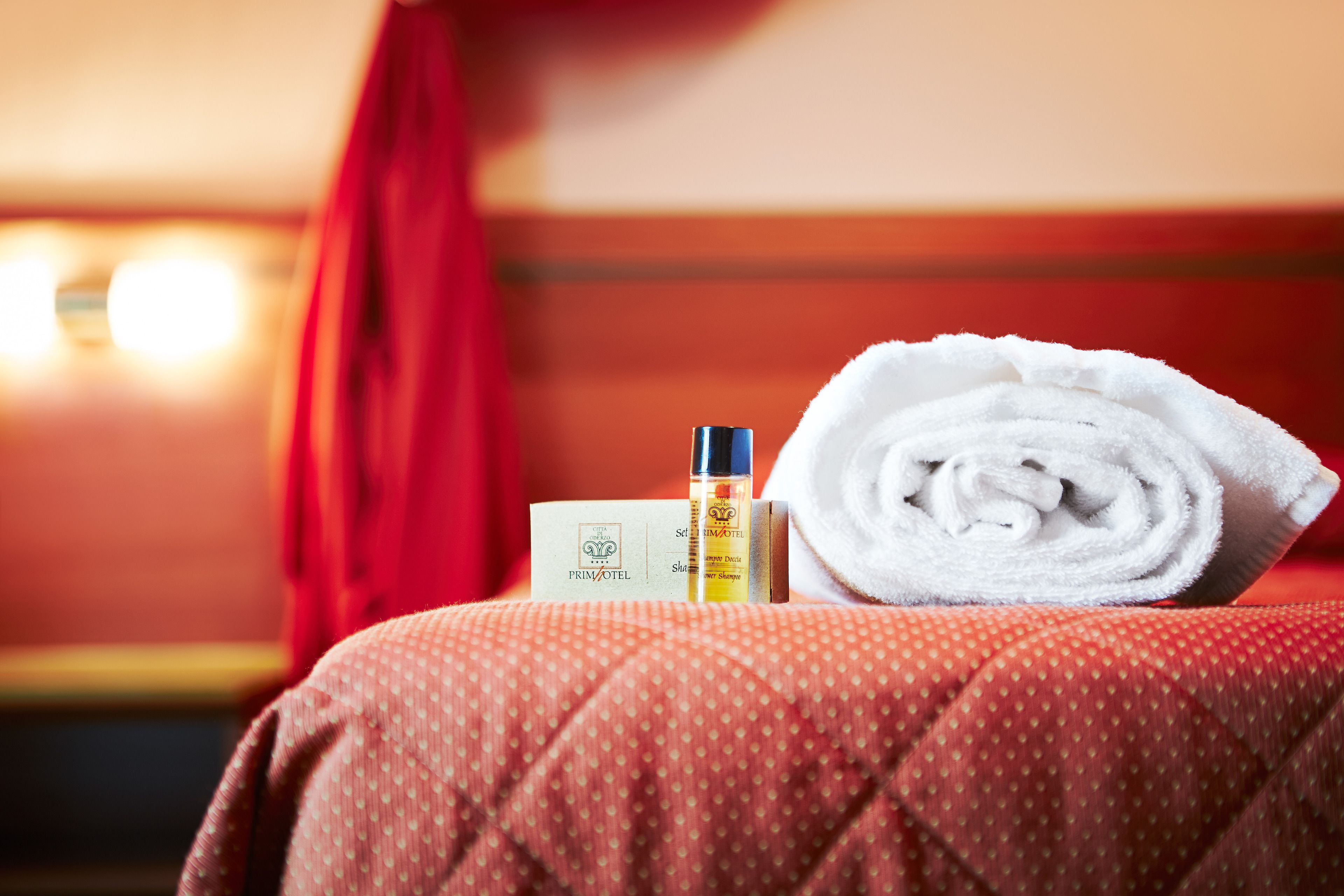 A neatly made bed with a rolled white towel, a bottle of lotion, and a card on a red bedspread in a hotel room.