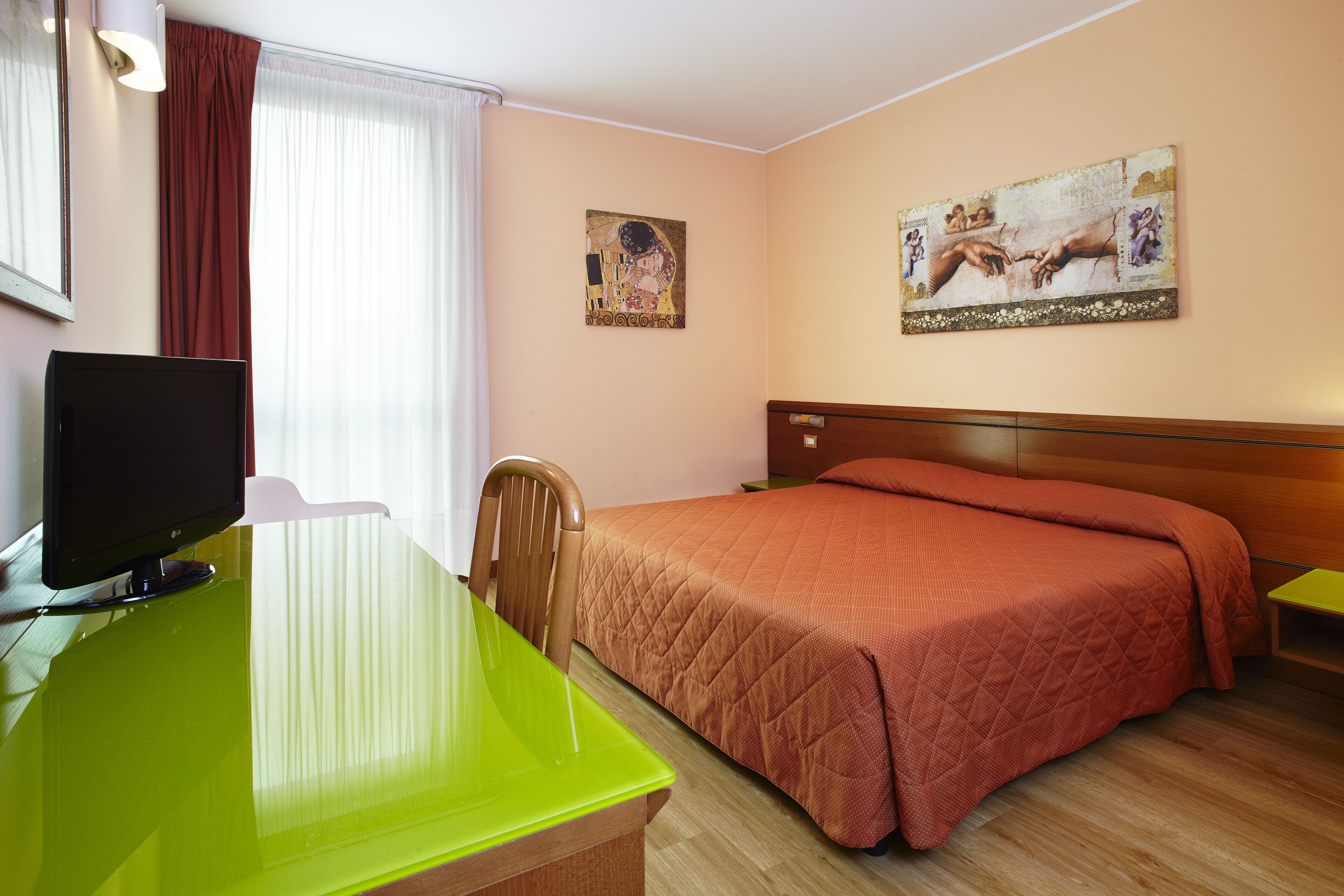 Una habitación de hotel con cama, escritorio, silla, televisor y obras de arte en las paredes.