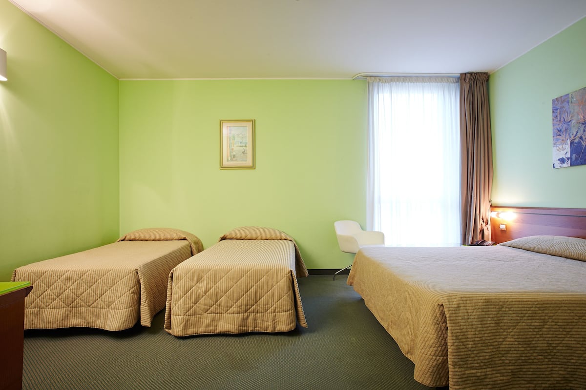 Quarto de hotel com duas camas de solteiro, uma escrivaninha, uma cadeira, um quadro na parede e uma janela com cortinas.