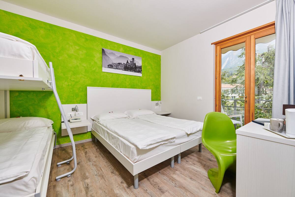 A hotel room with a bunk bed, a single bed, a desk with a chair, and a window with a view of buildings and trees.