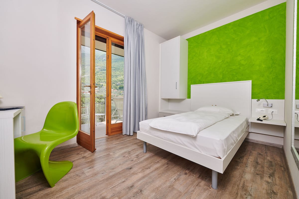 A bedroom with a bed, a green chair, a window with curtains, and a view of greenery outside.