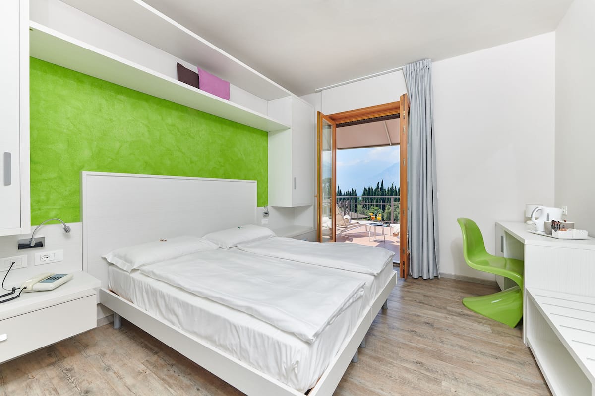 A modern hotel room with a large bed, a desk with a chair, and a balcony with a view of trees and mountains.