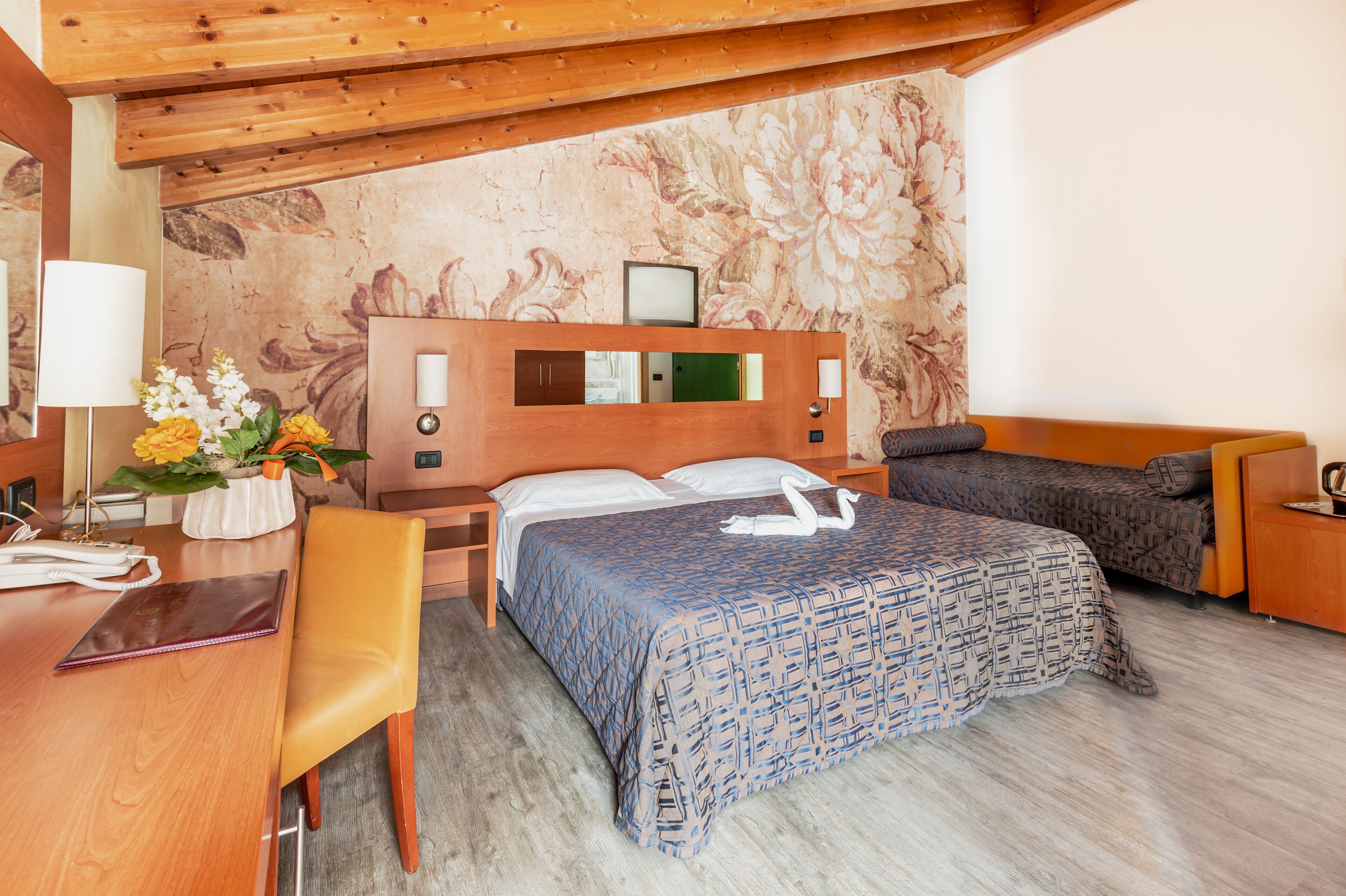 A hotel room with a wooden ceiling, a bed with patterned bedding, a desk with a chair, and a nightstand with a lamp and flowers.
