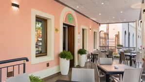 Un café interior con mesas y sillas, una planta grande en maceta y un cartel con un símbolo verde.