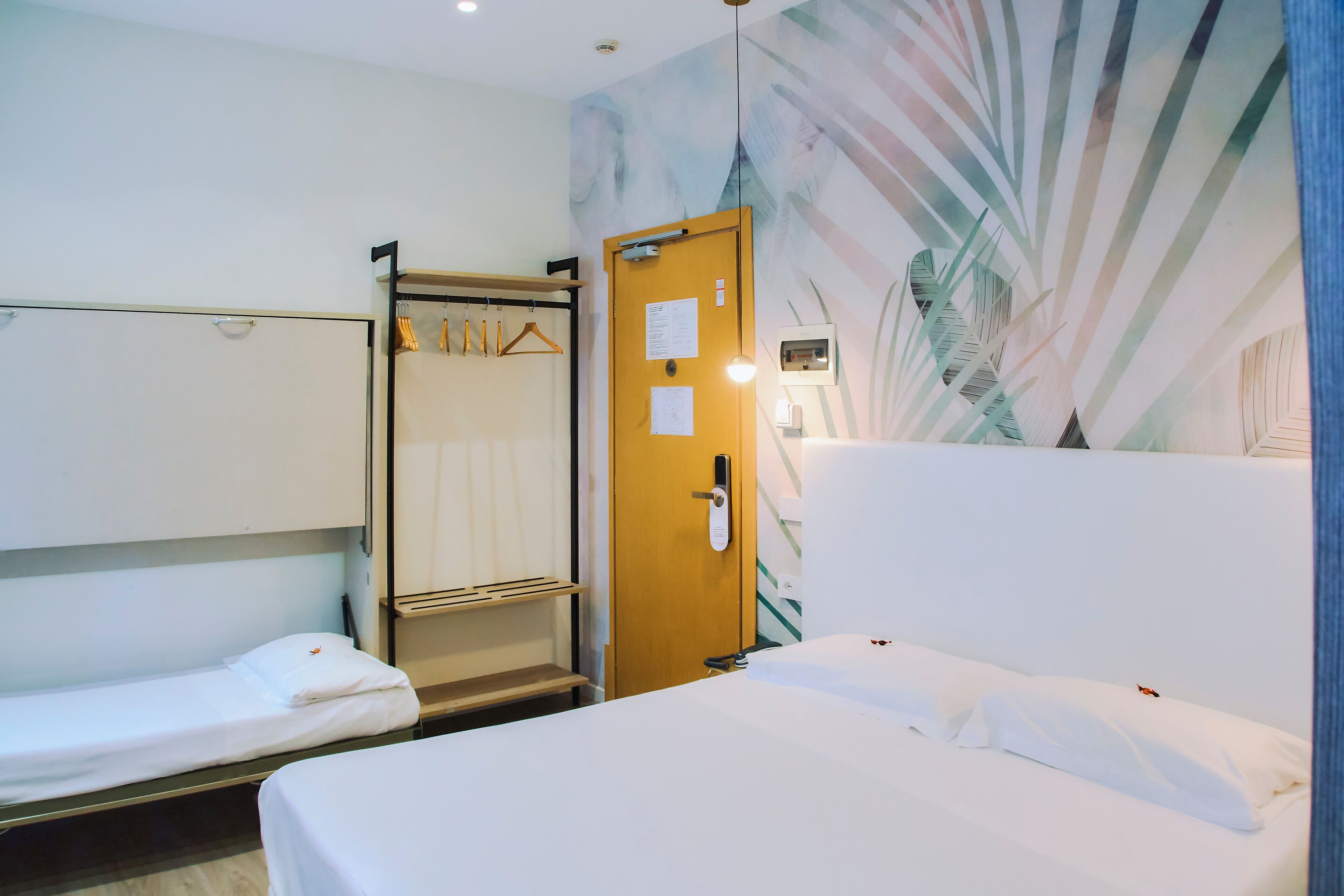 A hotel room with a bunk bed, a single bed, a wooden door with a phone, a coat rack, and a wall with a leaf pattern.