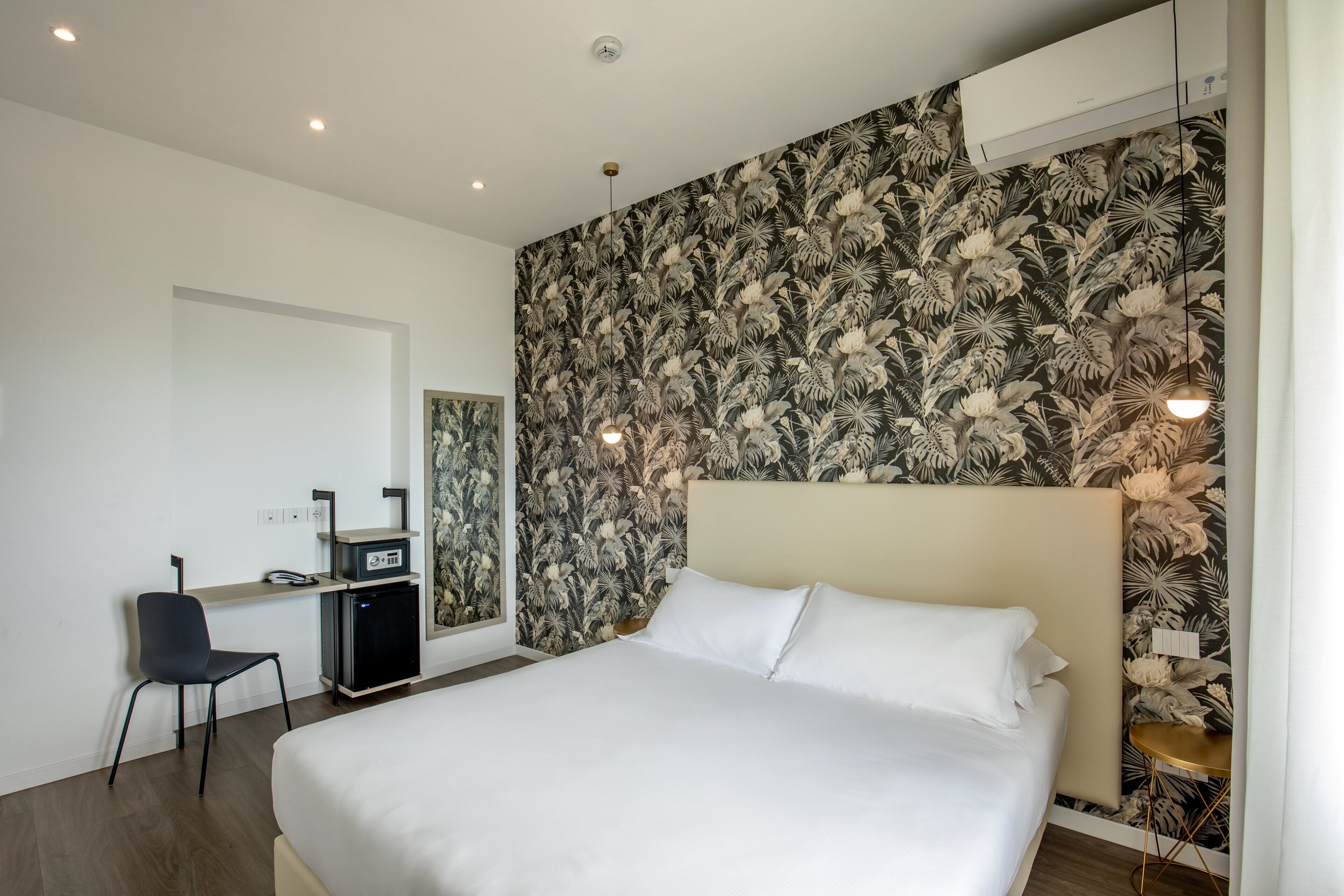 A modern hotel room with a large bed, a desk with a computer, a chair, and a small table.