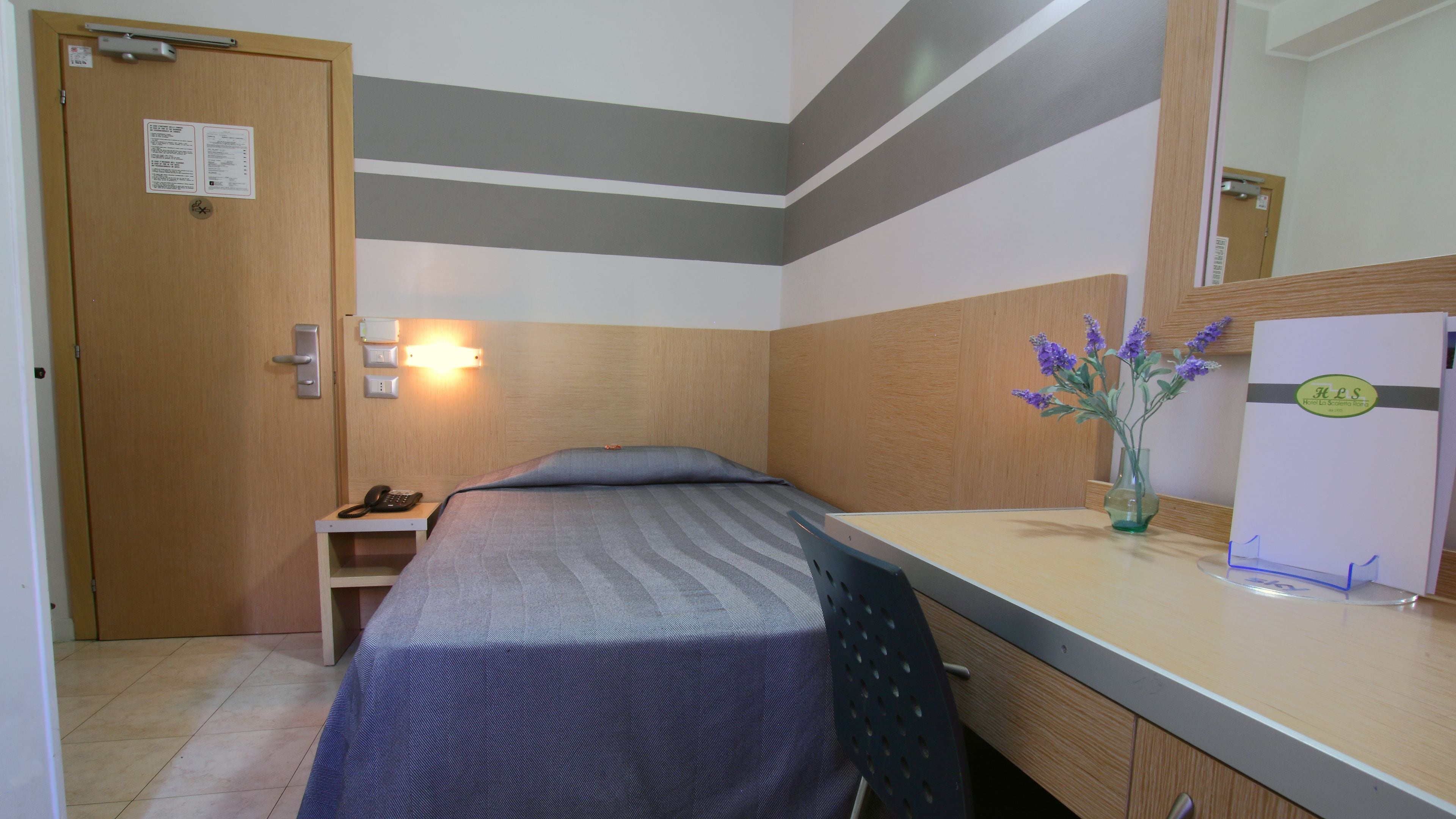 Una habitación de hotel con cama, mesita de noche, lámpara y un florero con flores.
