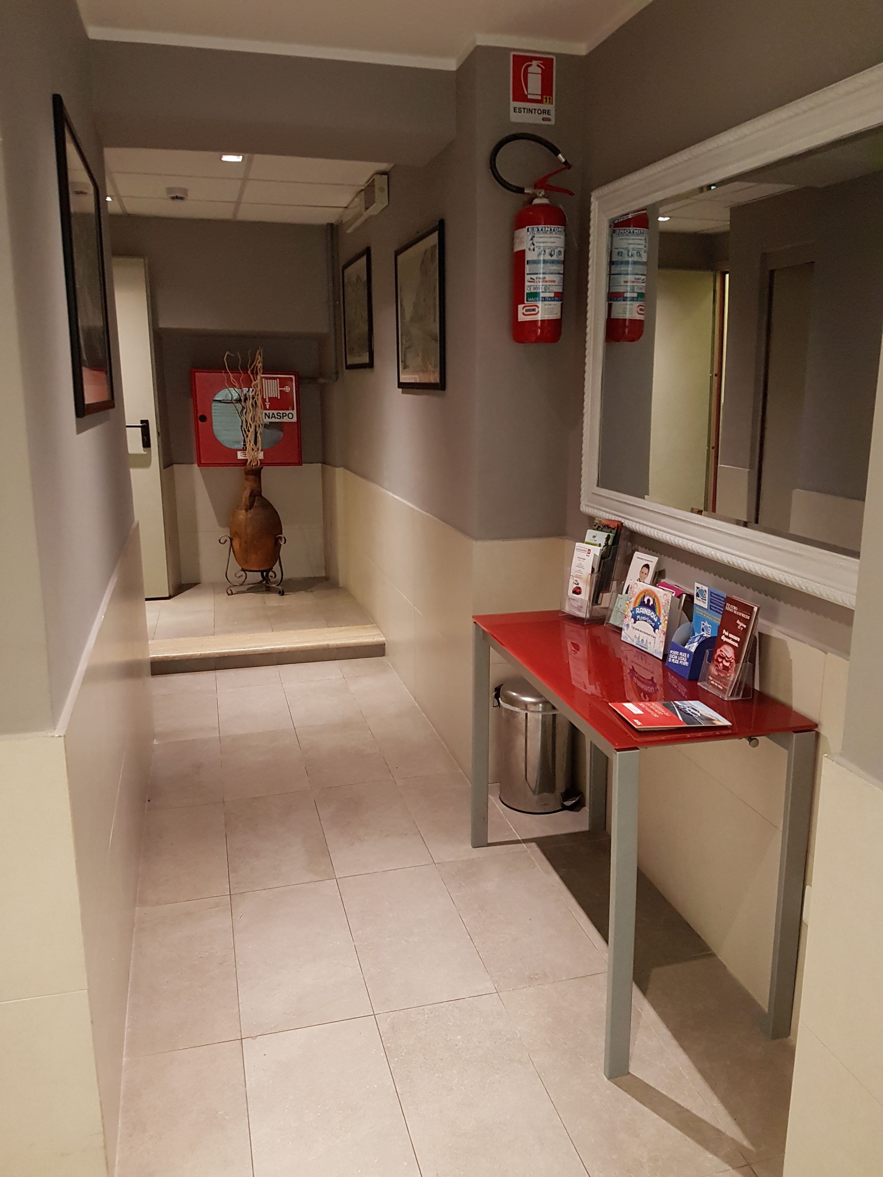 A hallway with a red reception desk, framed pictures on the walls, a fire extinguisher, and a trash bin.