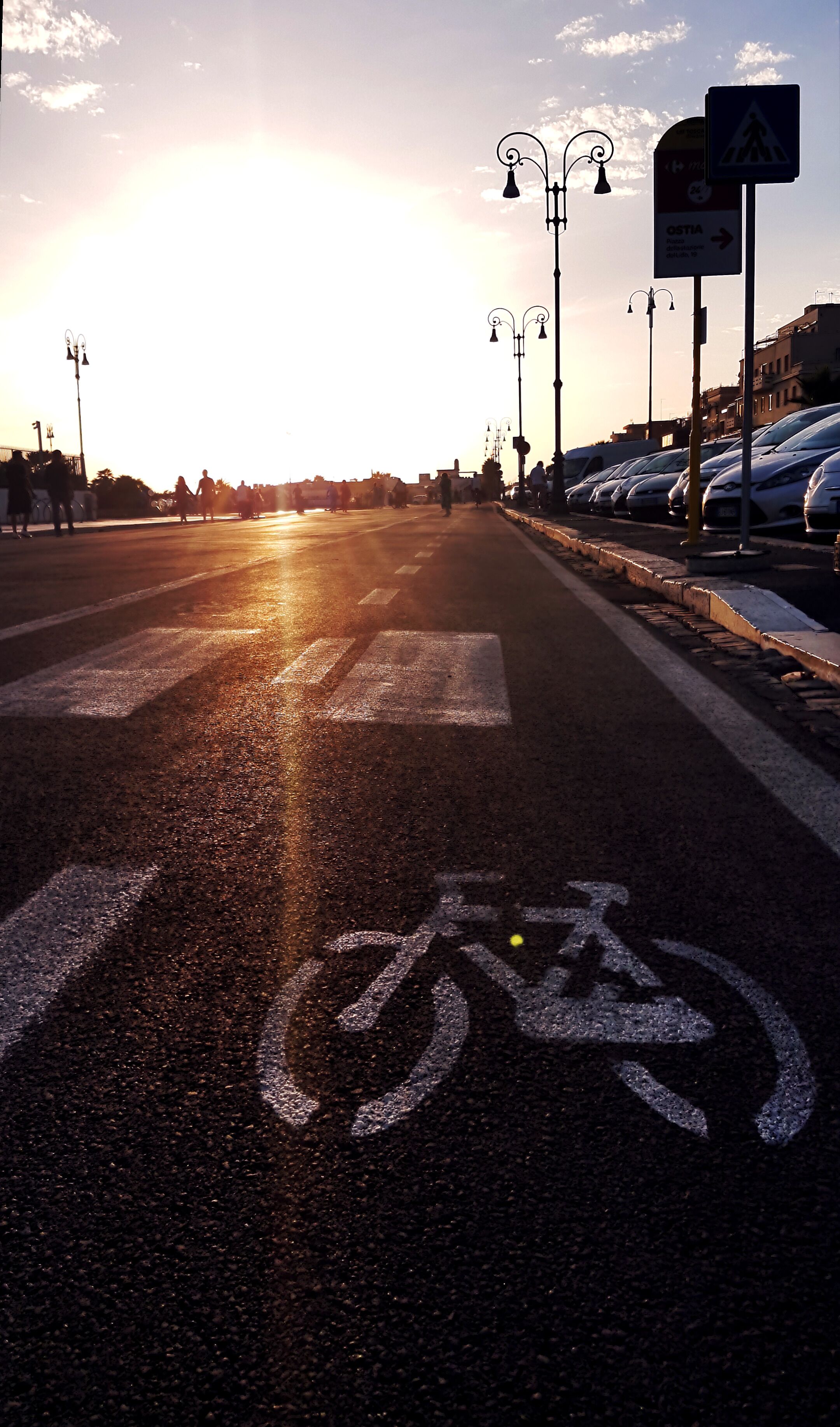 A road with a bicycle lane and pedestrian crossing, street lamps, and parked cars.