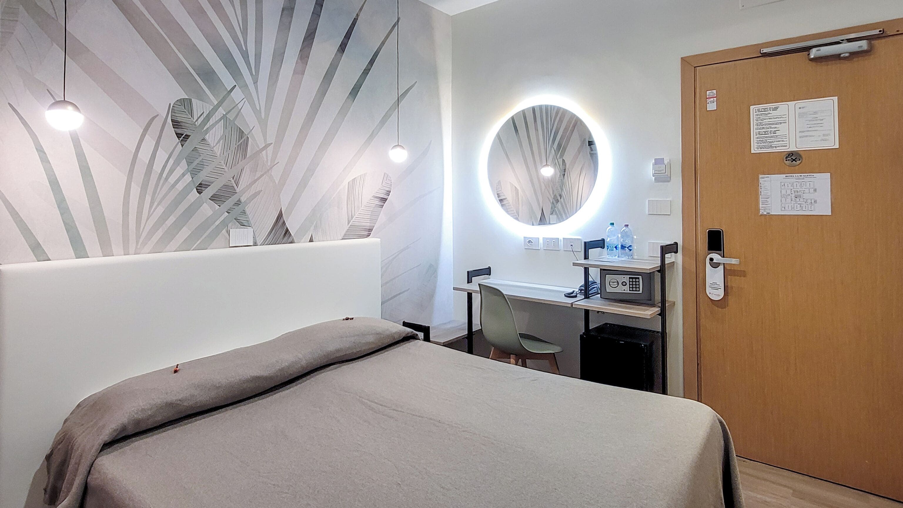 A hotel room with a bed, a desk with a computer, a mirror, and a door with a lock.