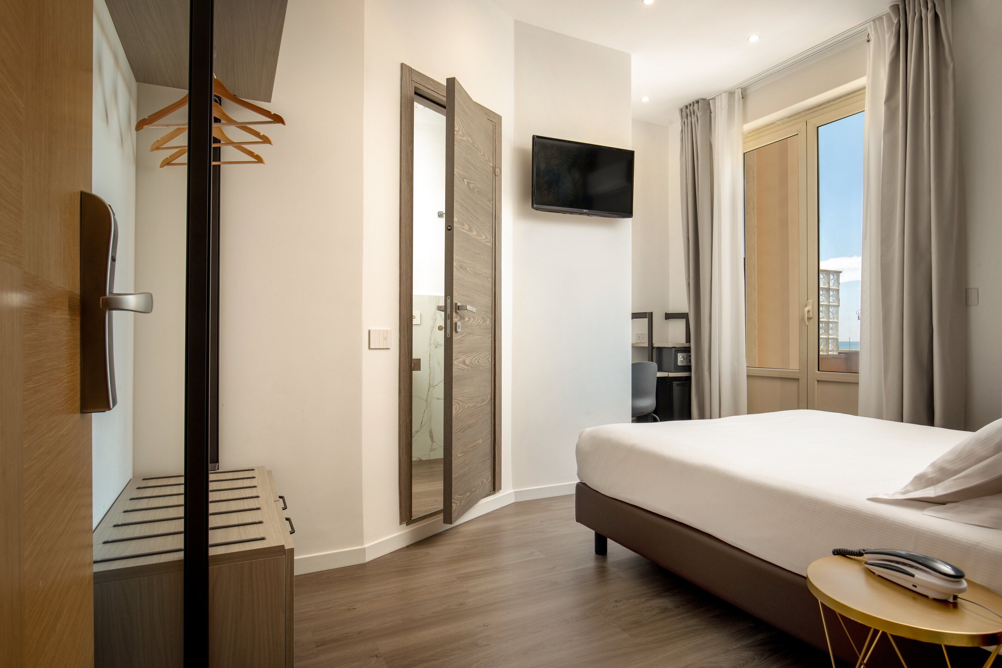 A modern hotel room with a bed, a TV, a desk, and a view of the city.