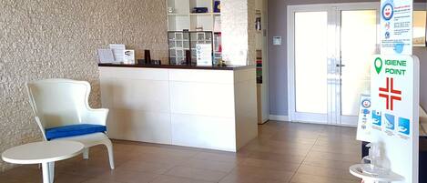 A reception area with a white reception desk, a white chair with a blue cushion, and a 'WiFi' sign. There is a 'Hotel Scalista' sign on the wall and a 'GIGIENE POINT' sign on the right.