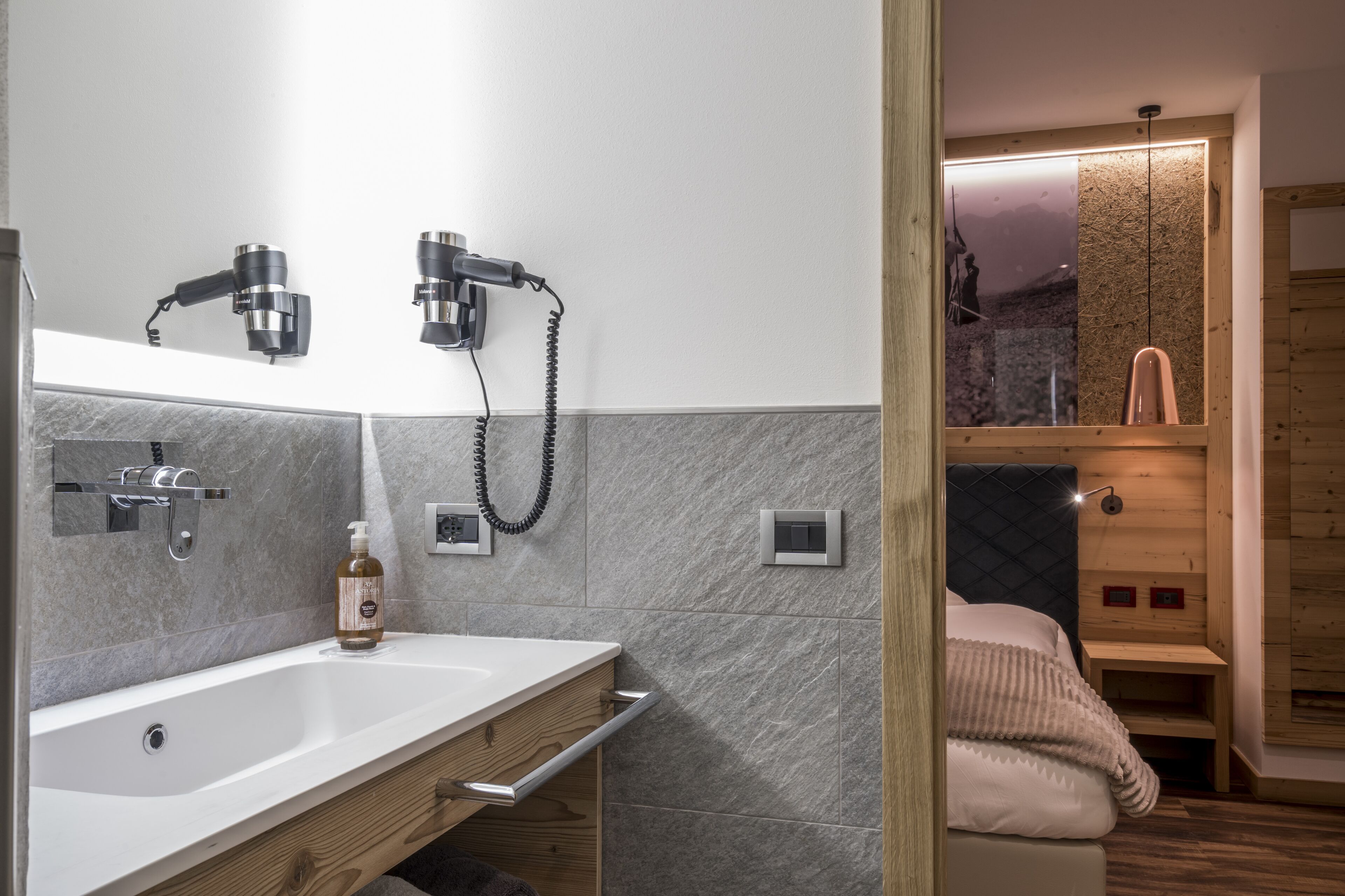 Un baño moderno con un lavamanos blanco, dos secadores de pelo montados en la pared, un mueble de tocador de madera y un espejo grande.